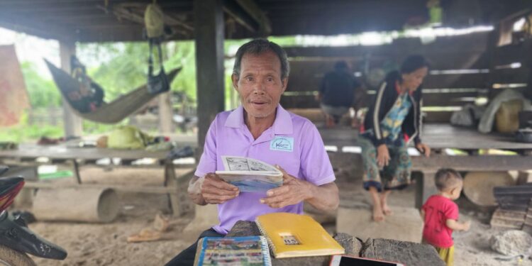 ប្រធានសហគមន៍នេសាទ លោក ខាន់ ប៊ុនទី ជាជនជាតិទំពួននៅភូមិកែងសាន់ ឃុំសេដា ស្រុកលំផាត់ ខេត្តរតនគិរី កំពុងអង្គុយកាន់សៀវភៅនៅក្រោមផ្ទះកាលពីថ្ងៃទី២៦ ខែកក្កដា ឆ្នាំ២០២៥។ (សាមន្ត ដារ៉ា)